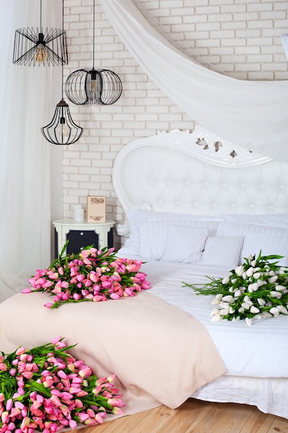 Romantic morning in a chic bedroom. A large bouquet of pink tulips lie on a white bed. Classic bedroom design. Brick white wall. Many unusual geometric lamps. Elegant luxury bedroom. White interior