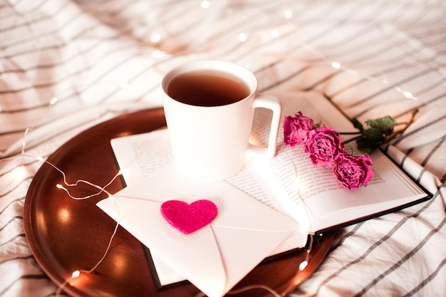 Romantic morning with cup of tea staying on open book with flowers in bed closeup. Valentines day.