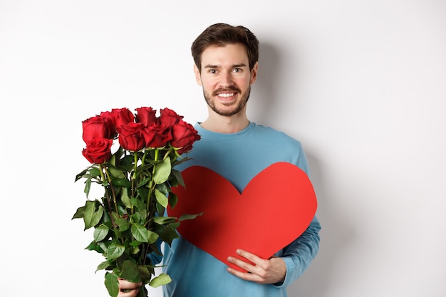 Romantic people. Handsome smiling man holding bouquet of roses and big red heart, going on Valentines day date with girlfriend, standing on white