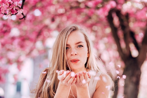 Photo romantic portrait of charming blonde on the wall of a blooming garden. tree with pink flowers. beauty blonde woman with long hair outdoors. cherry blossoms. close up portrait. sakura