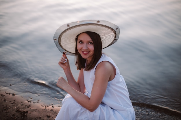 A romantic portrait of a laughing girl in a white sundress and a straw hat on a date on the beach by...