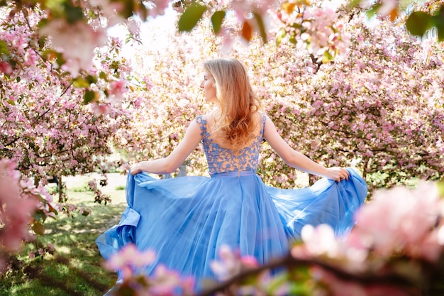 Romantic portrait of a runaway beautiful young girl in a long ultramarine blue dress of pink sakura in the garden