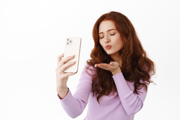 Романтичная глупая рыжая девушка посылает воздушный поцелуй в камеру смартфона, делает кокетливое селфи-фото, видеозвонок любовному партнеру в приложении для мобильного телефона, стоя на белом фоне