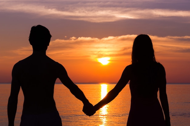 Романтическая сцена Дня святого Валентина молодой пары силуэтов, взявшись за руки у моря, глядя на красочный закат.
