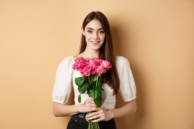 Romantic Valentines day concept. Beautiful young lady holding pink roses and smiling, standing happy on beige