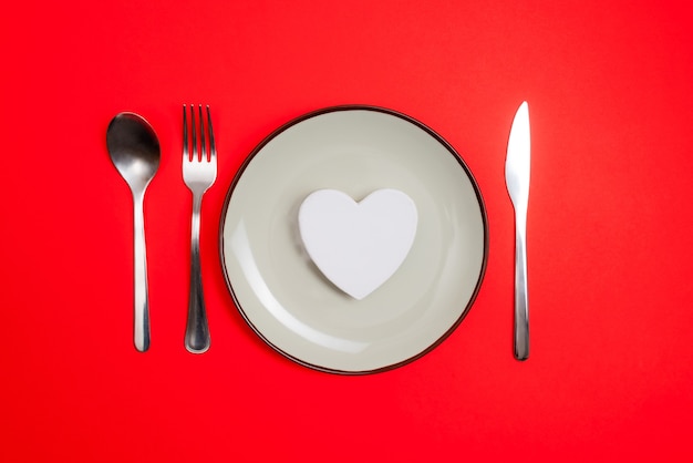 Romantic valentines day dinner idea concept. Heart on plate and silver wear on red background.