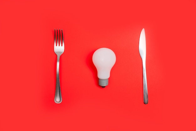 Romantic valentines day dinner idea concept. Light bulb on plate and silver wear on red background.