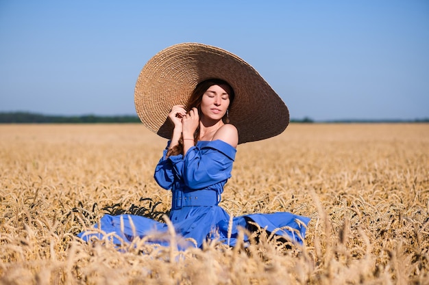 Romantic vintage lady in blue midi dress with belt and wide brimmed hat stand inside countryside golden wheat field