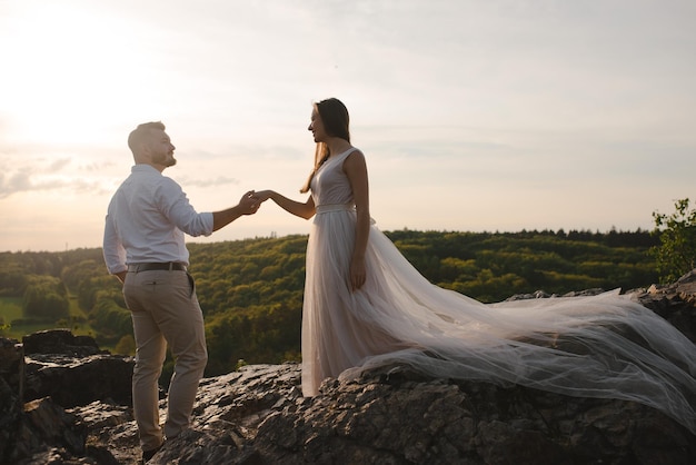 Romantic wedding couple standing on the rocks on sunset Bride woman in beautiful dress with long loop and stylish groom holding hands Sunset in green mountains