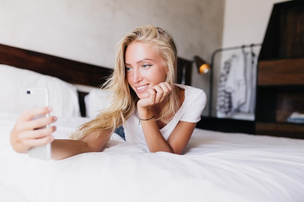 Romantic white girl making selfie in her bedroom Appealing fairhaired woman taking picture of herself while lying in bed