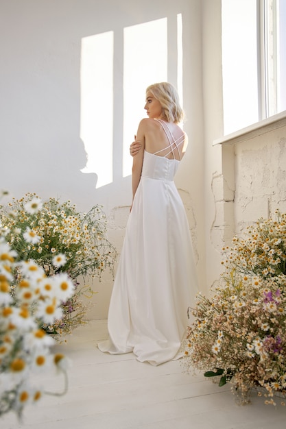 Фото Романтичная женщина в длинном белом свадебном платье стоит у окна в цветах ромашки