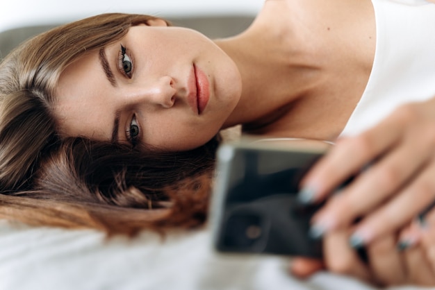 Photo romantic woman looking at mobile phone while laying on her bed. brunette young woman using cellphone at home. beautiful girl surfing the net on smartphone and relaxing
