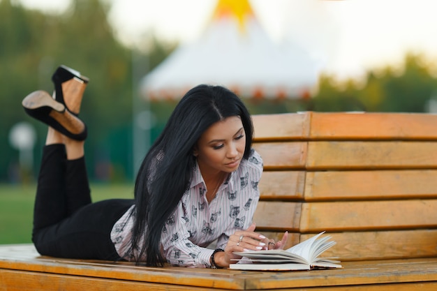 Romantica giovane ragazza bruna in abiti eleganti che legge un libro sdraiato su una panca di legno nel parco in una calda giornata estiva