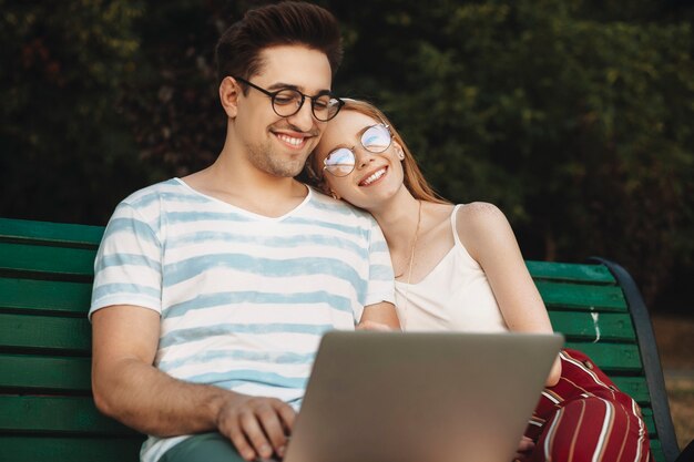 야외 웃 고 벤치에 앉아 로맨틱 젊은 부부. 빨간 머리 소녀가 웃고 그에게 머리를 기울이고있는 동안 노트북에 소셜 미디어를하는 남성 동영상 블로거.