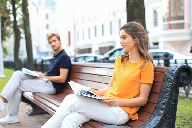 Romantic young couple in summer clothes smiling and reading books together while sitting on bench in city street.