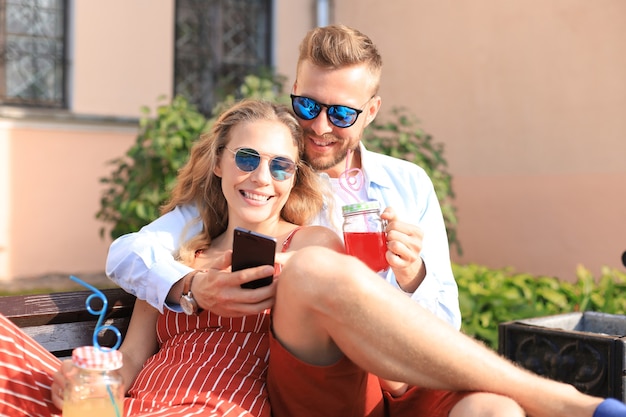 Romantic young couple in summer clothes smiling and taking selfie while sitting on bench in city street.