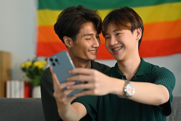 LGBT와 사랑 개념을 배경으로 거실 무지개 깃발에 스마트폰으로 셀카를 찍는 로맨틱한 젊은 게이 커플