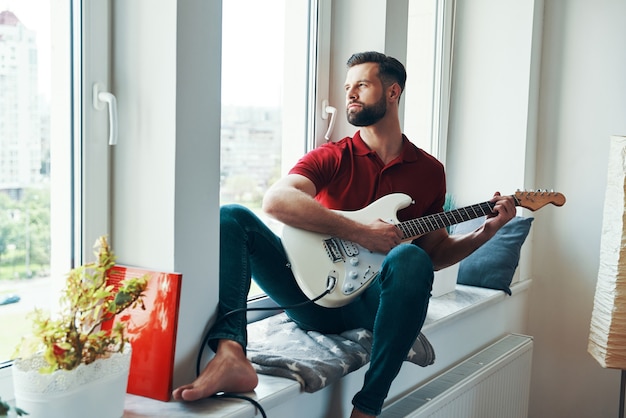 Giovane romantico in abbigliamento casual che suona la chitarra mentre è seduto sul davanzale della finestra