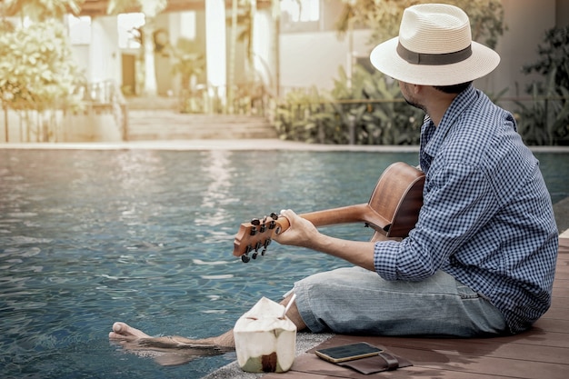 Romantic young man sitting on the pool at sunset with playing the guitar