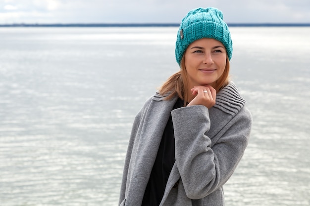 Romantic young woman on blue natural sea background outdoors. Portrait of a beautiful young brunette woman on the beach, she has long hair and wears coat and knitting hat