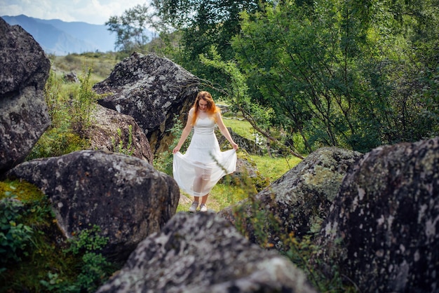 La giovane donna romantica cammina lungo un sentiero tra enormi pietre grigie. bruna dai capelli lunghi in abito bianco sullo sfondo di un pittoresco paesaggio di montagna.