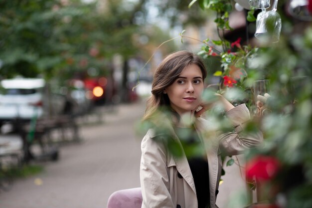 ロマンチックな若い女性は、朝、ストリートカフェに座ってベージュのコートを着ています。緑と花の外飾り。テキスト用のスペース