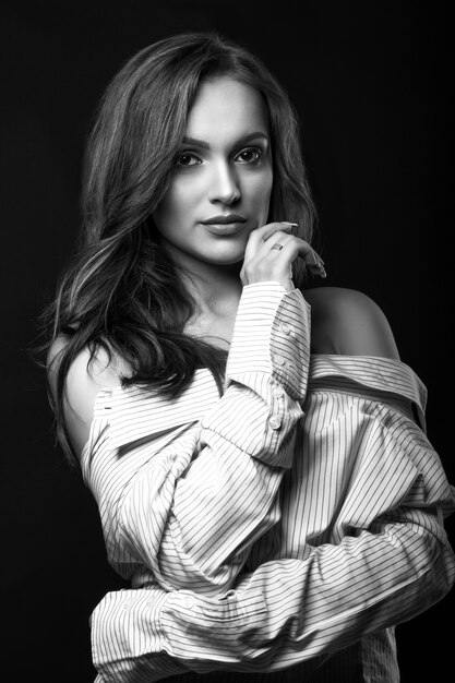 Romantic young woman with natural makeup posing in shirt at studio with dramatic light. Black and white portrait