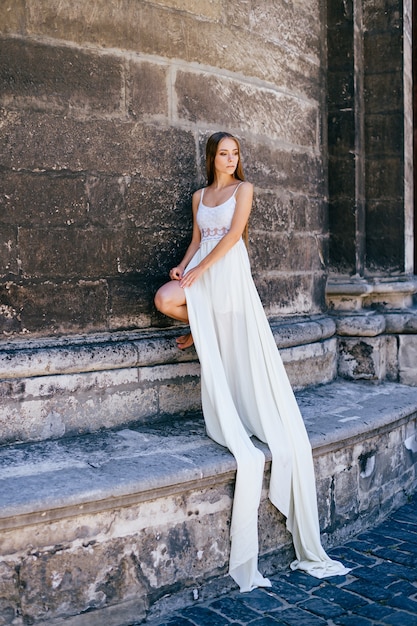 Foto romantisch elegant meisje in lange witte jurk poseren over oude stenen muur