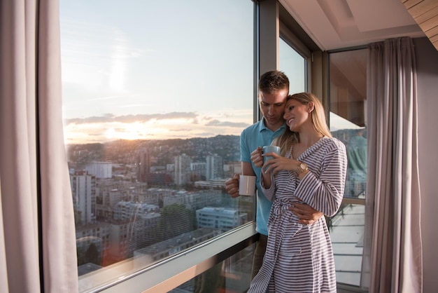 romantisch gelukkig jong stel dat 's avonds geniet van koffie en een prachtig zonsonderganglandschap van de stad terwijl ze bij het raam staan