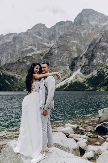 Romantisch huwelijkspaar in liefde die zich op de steenachtige kust van het Sea Eye-meer in Polen bevinden. Schilderachtig uitzicht op de bergen. De bruid en bruidegom. Morskie Oko. Tatra-gebergte.