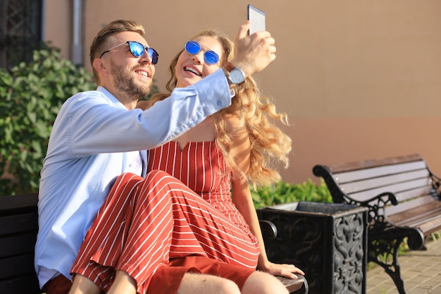 Romantisch jong stel in zomerkleren glimlachend en selfie nemend terwijl ze op een bankje in de straat van de stad zitten.