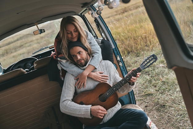 Romantisch liedje spelen. Knappe jongeman die gitaar speelt voor zijn mooie vriendin terwijl hij in een minibusje in retrostijl zit