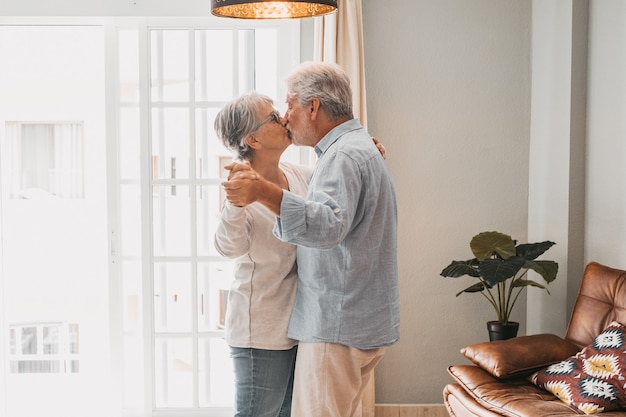 Romantisch liefdevol senior koppel hand in hand genietend van samen dansen terwijl ze elkaar kussen in de woonkamer van het huis, paar zoenen en omhelzen terwijl ze elkaars hand vasthouden thuis