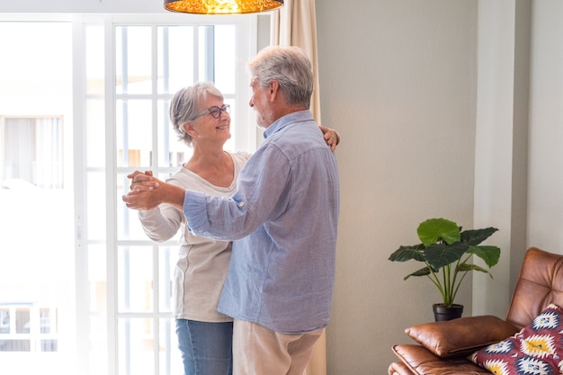 Romantisch liefdevol senior paar hand in hand genieten van samen dansen in de woonkamer van het huis, bejaarde gelukkige paar vieren door te dansen in de woonkamer.