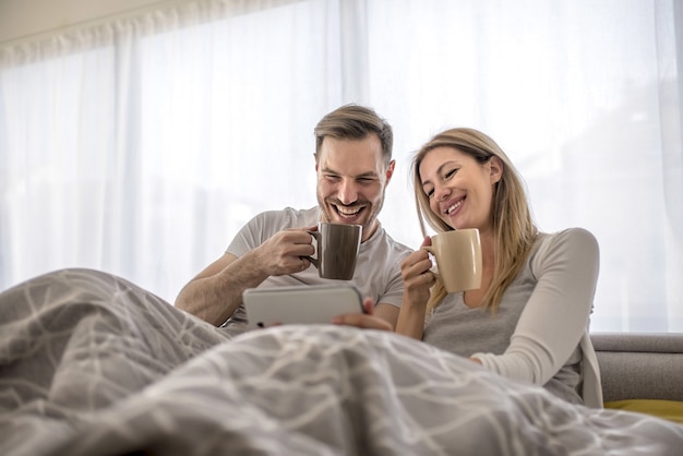 Romantisch paar dat in het bed ligt en koffie drinkt en iets op hun telefoon bekijkt