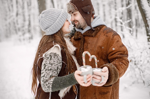 Romantisch paar dat in het winterbos staat en twee kopjes met cacaodrank vasthoudt