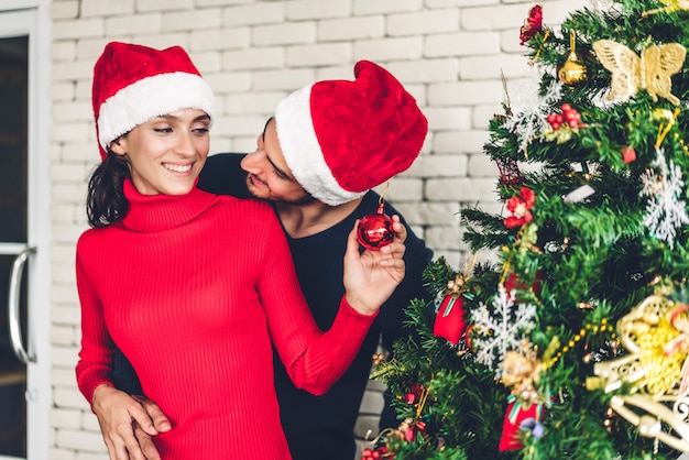 Romantisch zoet paar in santahoeden die Kerstmisboom verfraaien en terwijl het vieren van nieuwe jaarvooravond en het genieten van samen tijd doorbrengen glimlachen