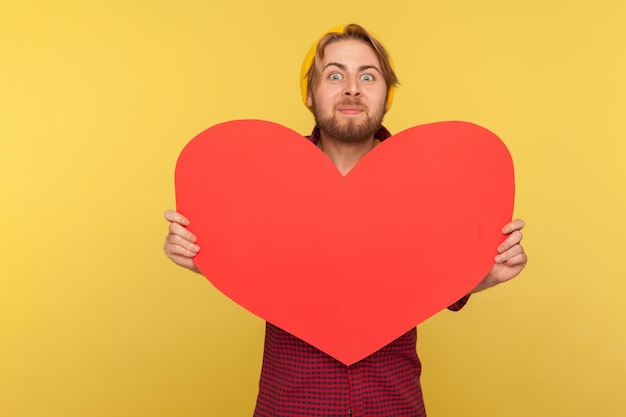 Romantische man die een groot gelezen hart vasthoudt en met opwinding naar de camera kijkt, die gevoelens van liefde en genegenheid demonstreert met een enorme wenskaart op Valentijnsdag. studio-opname geïsoleerd op gele achtergrond