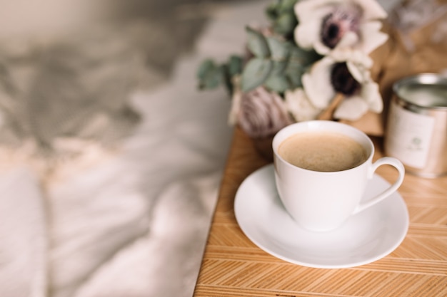 Romantische ochtend. Houten salontafel met bloemen op bed met plaid, koffiekopje, bloemen en kaarsen. Lila rozen met eucalyptus en anemonen. Grijze tinten interieur.