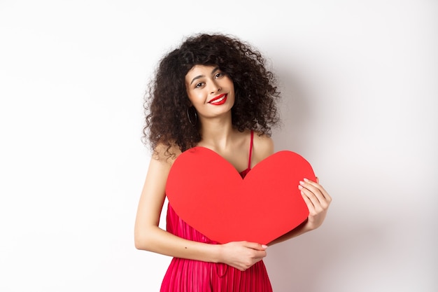 Romantische tedere vrouw met krullend haar, groot rood hart knuffelen en glimlachen, kijken met liefde, staande tegen een witte achtergrond.