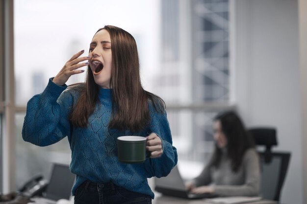 Rommelige en slapeloze jonge witte kraag Turkse vrouw in een modern kantoor met haar collega's Ze geeuwt en houdt een kopje koffie