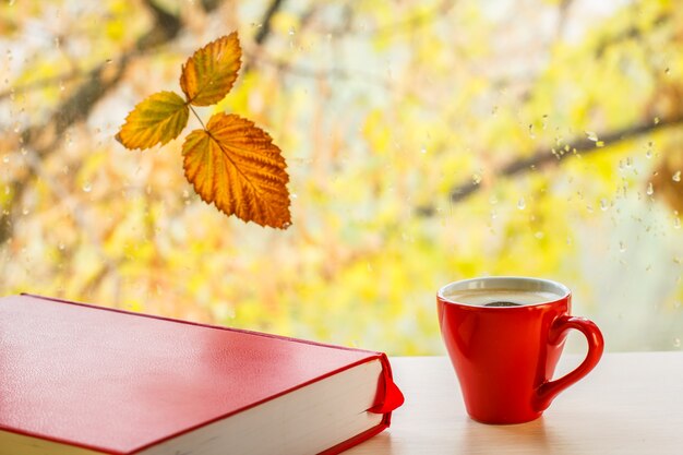 Rood boek, kopje koffie en droog blad op vensterglas met waterdruppels in de wazige natuurlijke achtergrond. Gevallen blad en regendruppels op een ruit met herfstbomen op de achtergrond.