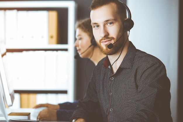 Roodbaard zakenman praten door headset in de buurt van zijn vrouwelijke collega zittend in een zonnig kantoor. Diverse mensengroep in callcenter.