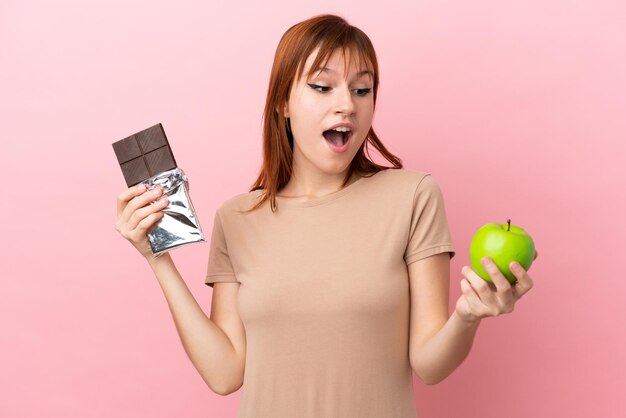 Roodharig meisje geïsoleerd op roze achtergrond met een chocoladetablet in de ene hand en een appel in de andere