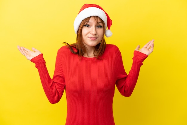 Roodharig meisje met kerstmuts geïsoleerd op gele achtergrond met twijfels tijdens het opsteken van de handen