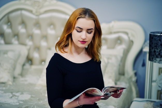 Roodharig meisje op zwarte kledingszitting op bed en het lezen van modetijdschrift