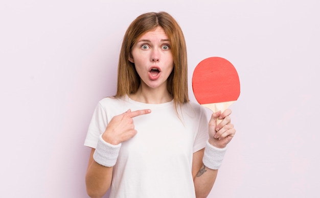 Roodharig mooi meisje dat geschokt en verrast kijkt met wijd open mond wijzend naar zelf pingpong concept