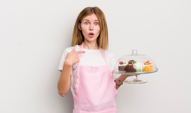 Foto roodharig mooi meisje dat geschokt en verrast kijkt met wijd open mond wijzend naar zelfgemaakt taartenconcept