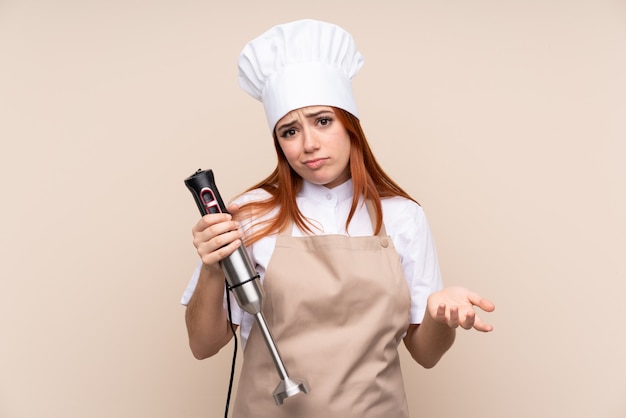 Roodharig tienermeisje die handmixer over geïsoleerde muur gebruiken die twijfelsgebaar maken terwijl het opheffen van de schouders