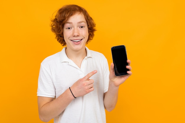 Roodharige aantrekkelijke knappe jongen in een wit T-shirt met een telefoon met een mockup op een gele achtergrond